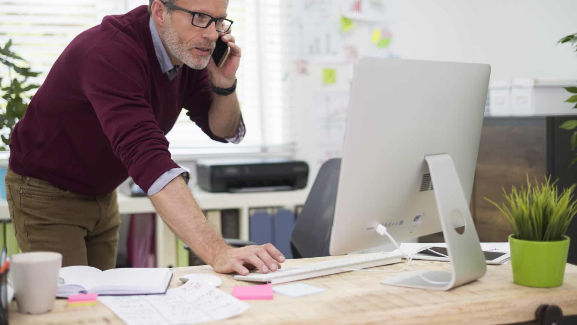 Mann am Schreibtisch mit Telefon am Ohr, Rechner, Notizbuch und Haftzetteln: Wer sein CRM automatisieren will, sucht Kundendaten sonst an vielen Orten